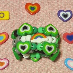 May include: A beaded bracelet with green heart-shaped beads and a rainbow charm. Surrounding the bracelet are heart-shaped charms in various colors, including rainbow, purple, and pink. A yellow rectangle with a purple circle is also present.