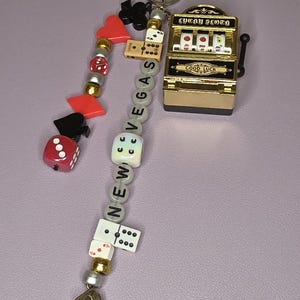 May include: A keychain featuring a miniature gold slot machine, dice, playing card charms, and beads spelling "NEW VEGAS." The keychain includes a metal ring for attachment.
