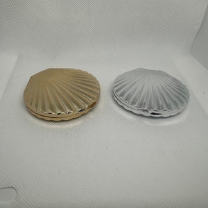 Puede incluir: Dos compactos decorativos pequeños en forma de concha. Uno es dorado y el otro es plateado.