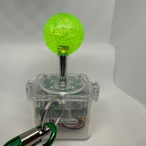 May include: A clear plastic arcade joystick with a green, bubble-textured ball on top. A green carabiner clip is attached to the base.
