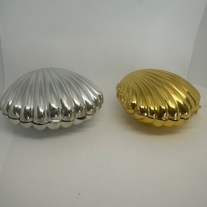 May include: Two decorative seashell-shaped boxes, one silver and one gold. The boxes are closed and have a smooth, shiny finish.