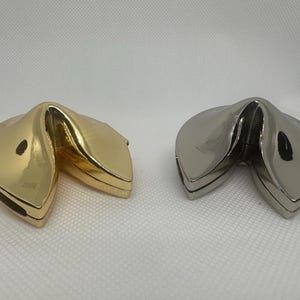 May include: Two fortune cookie shaped boxes, one gold and one silver, are open and laying on a white surface.