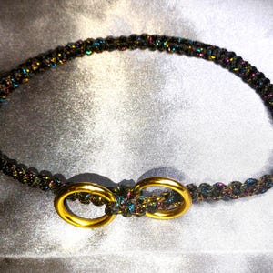 May include: A black and iridescent beaded belt with gold-colored double-ring clasp. The belt is made of tightly woven beads that shimmer with colors of blue, purple, and green. The clasp is a bright gold color.