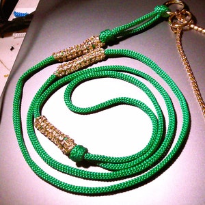 May include: A bright green braided cord lanyard featuring three cylindrical woven accents in a metallic gold tone. The cord is secured with knots and terminates in a gold-toned metal ring at one end, with a matching gold-toned chain attached.