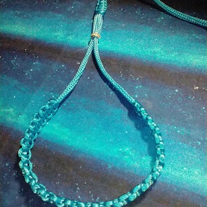 May include: A teal blue paracord lanyard with a gold metal bead. The lanyard is made of braided rope and has a loop at the end.