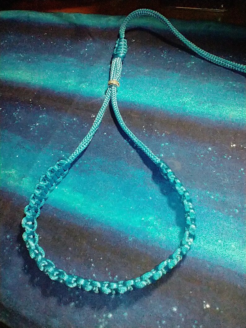 May include: A teal blue paracord lanyard with a gold metal bead. The lanyard is made of braided rope and has a loop at the end.