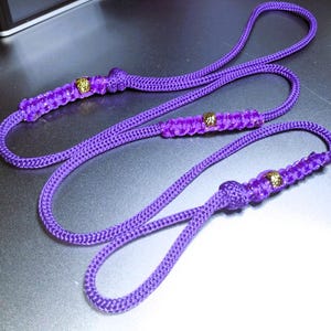 May include: A long purple cord made of braided nylon rope featuring three distinct sections of intricate knotwork. Each knotted segment is accented with a small, textured gold-colored metal bead. The cord is flexible and forms several loops.