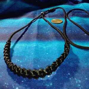 May include: A black braided cord necklace with a simple clasp. The necklace is displayed on a blue and black patterned fabric.