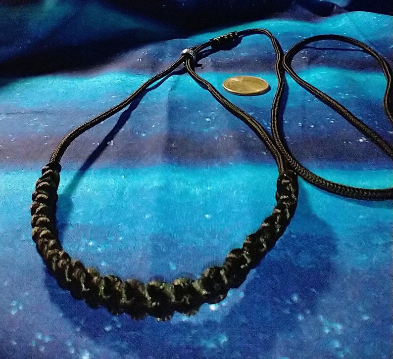 May include: A black braided cord necklace with a simple clasp. The necklace is displayed on a blue and black patterned fabric.