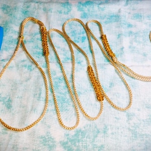 May include: A long, braided gold-colored cord featuring three cylindrical, amber-toned beaded sections. The cord is displayed next to a blue Sharpie marker for scale, showing the length of the cord relative to the approximately 5.5-inch marker.