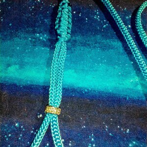 May include: A teal blue cord with a gold bead and a knot at the top. The cord is on a blue and black fabric background.