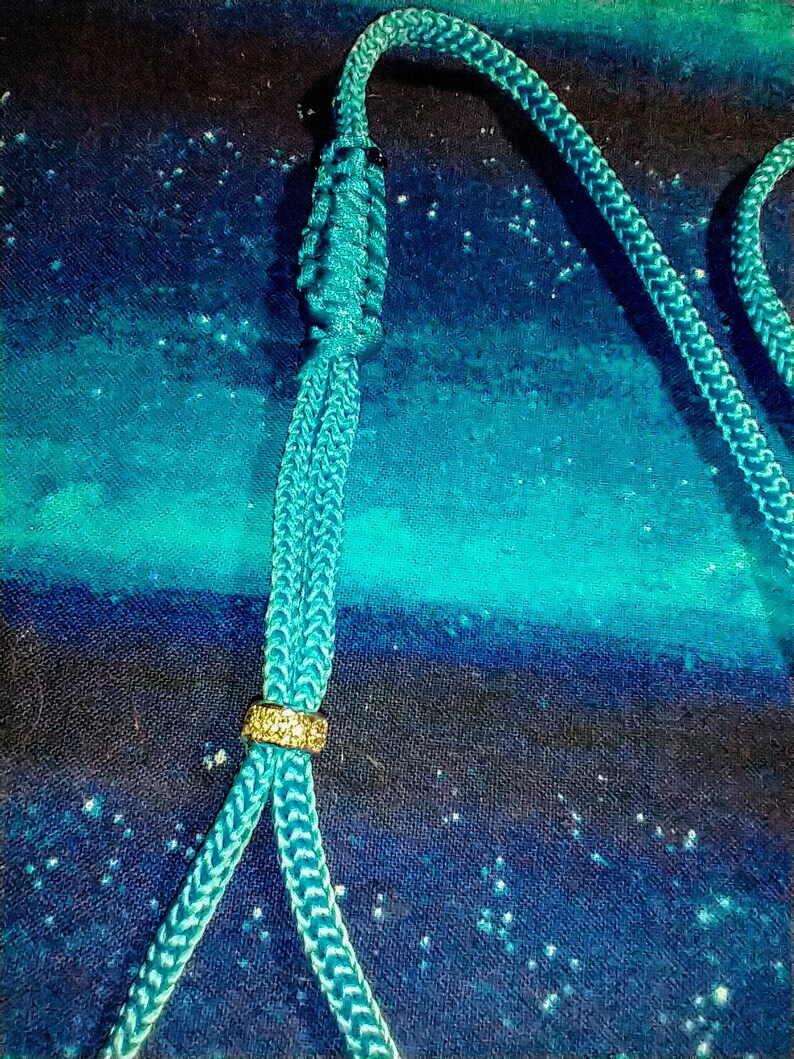 May include: A teal blue cord with a gold bead and a knot at the top. The cord is on a blue and black fabric background.