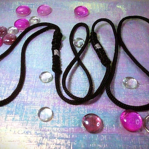 May include: A black cord lanyard featuring two adjustable sliding knots with silver-toned metal accents and a circular metal ring at one end. The cord is constructed from a braided material and is arranged in a series of loops across the frame.