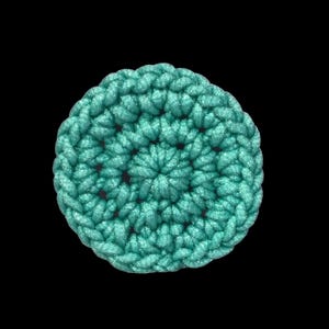 May include: A round, turquoise crocheted item against a black background. The item is made of thick yarn, with a spiral pattern from the center outward. The texture is visible, with each stitch clearly defined. The overall shape is circular.
