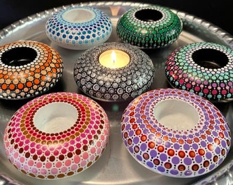 May include: Seven tealight candle holders, each painted with a different mandala design in various colors, including orange, green, blue, pink, purple, and gray. The candle holders are arranged in a circle on a silver tray.