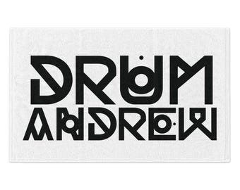 以下が含まれることがあります： 白地の長方形タオル。黒の太字で「DRUM ANDREW」と書かれています。文字はタオルの中央に配置され、「DRUM」が「ANDREW」の上にあります。タオルは柔らかく、質感のある外観をしています。