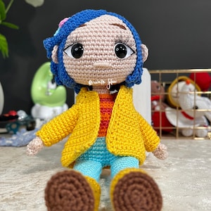 May include: A crocheted doll with blue hair, a yellow jacket, blue pants, and brown shoes. The doll has large brown eyes and a pink face. The doll is sitting on a white surface.