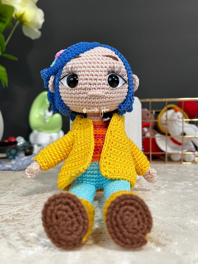 May include: A crocheted doll with blue hair, a yellow jacket, blue pants, and brown shoes. The doll has large brown eyes and a pink face. The doll is sitting on a white surface.
