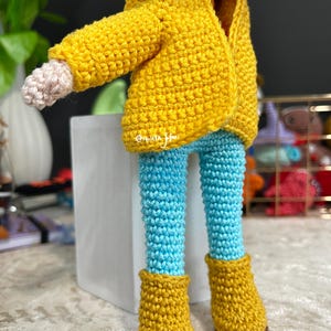 May include: A crocheted doll wearing a yellow jacket, blue pants, and yellow boots. The doll has blue hair and a crocheted face.