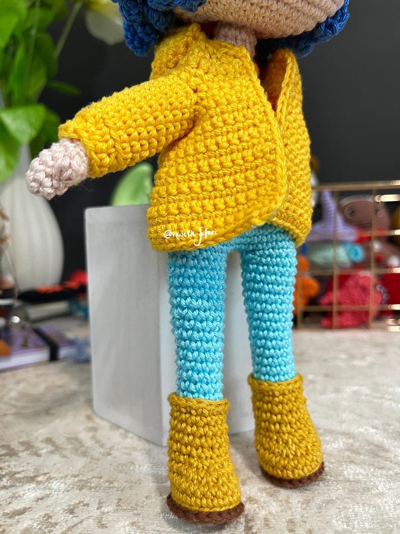 May include: A crocheted doll wearing a yellow jacket, blue pants, and yellow boots. The doll has blue hair and a crocheted face.