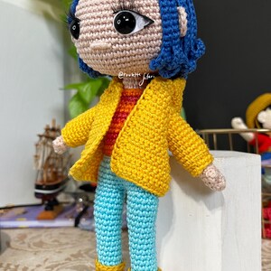 May include: Crocheted doll with blue hair, wearing a yellow jacket, blue jeans, and yellow boots. The doll is standing on a white block.