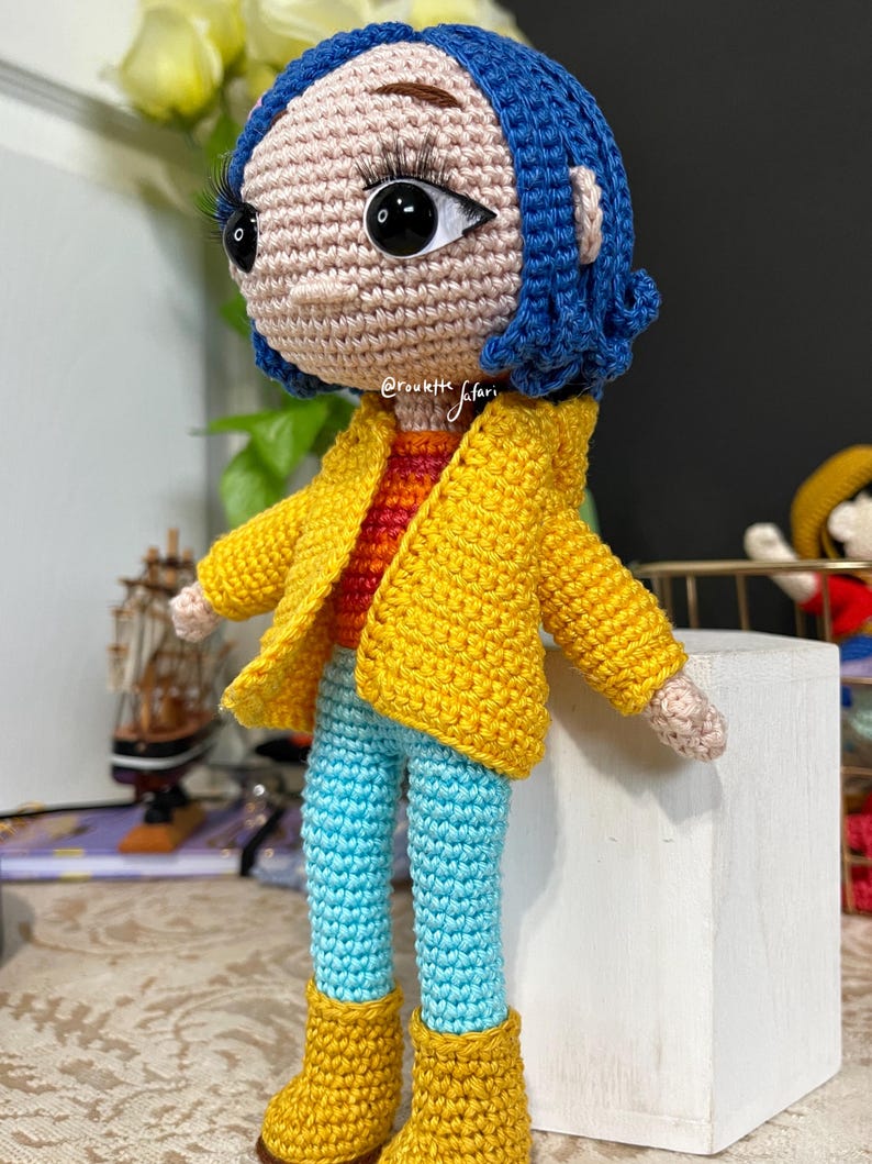 May include: Crocheted doll with blue hair, wearing a yellow jacket, blue jeans, and yellow boots. The doll is standing on a white block.