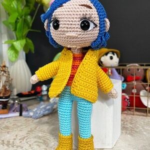 May include: A crocheted doll with blue hair, a yellow jacket, blue pants, and yellow boots. The doll has a pink flower in its hair and large brown eyes.