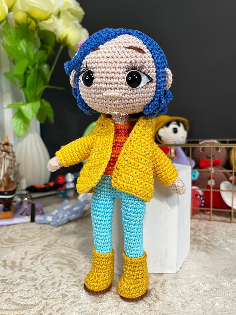 May include: A crocheted doll with blue hair, a yellow jacket, blue pants, and yellow boots. The doll has a pink flower in its hair and large brown eyes.