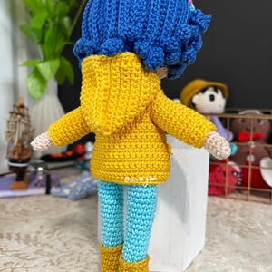 May include: A crocheted doll wearing a yellow raincoat, blue pants, and yellow boots. The doll has blue hair and a pink flower.