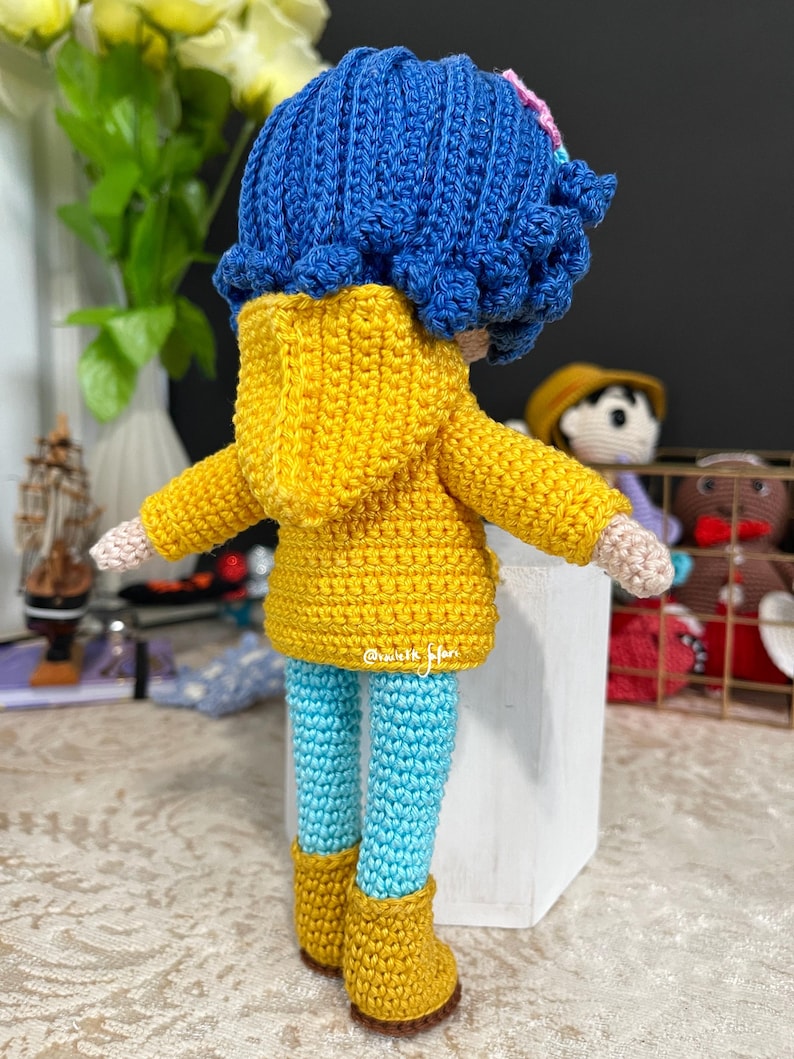 May include: A crocheted doll wearing a yellow raincoat, blue pants, and yellow boots. The doll has blue hair and a pink flower.