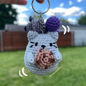 May include: A gray crocheted cat keychain holding a brown crocheted cookie. The keychain also features a smaller gray crocheted cat and a purple crocheted ball. The keychain is hanging from a silver key ring.