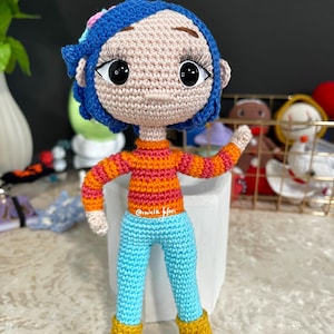 May include: A crocheted doll with blue hair, orange and pink striped shirt, blue pants, and yellow boots. The doll is standing on a white surface.