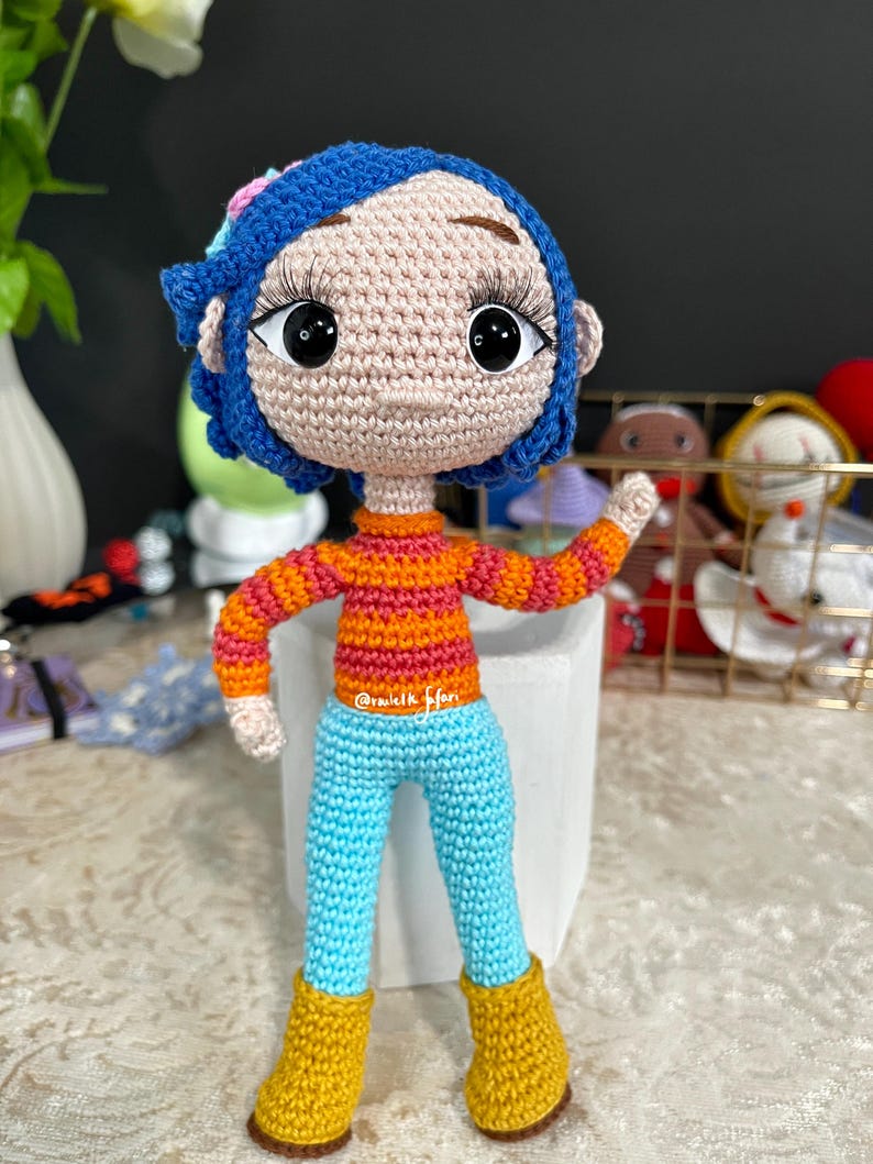 May include: A crocheted doll with blue hair, orange and pink striped shirt, blue pants, and yellow boots. The doll is standing on a white surface.