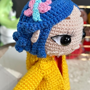 May include: A crocheted doll with blue hair and a yellow jacket. The doll has a pink and blue butterfly on its head. The doll's eyes are black and white.