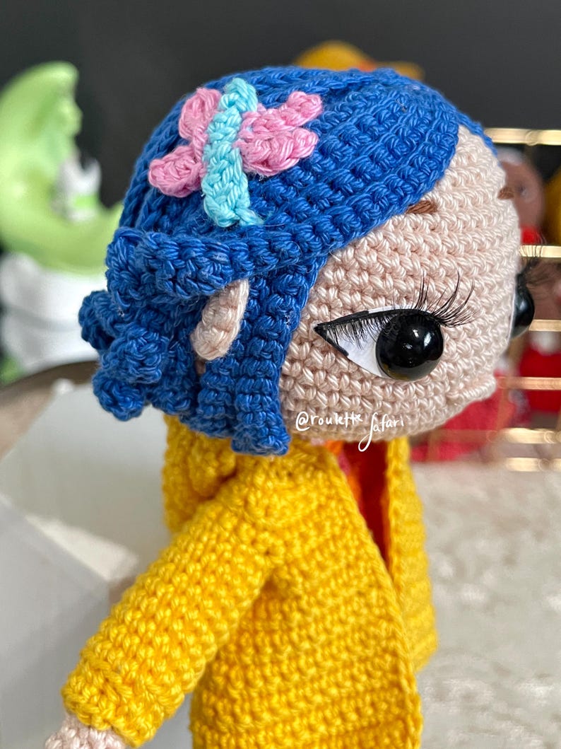 May include: A crocheted doll with blue hair and a yellow jacket. The doll has a pink and blue butterfly on its head. The doll's eyes are black and white.