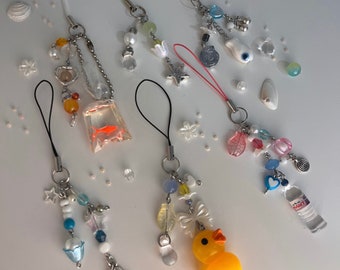 May include: A collection of colorful phone charms featuring various designs, including a fish, a duck, a heart, a star, and a bottle of water. The charms are attached to phone cases with strings and beads.