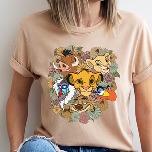 May include: A beige t-shirt with a graphic print of the Lion King characters Simba, Nala, Pumbaa, Timon, and Zazu surrounded by flowers.