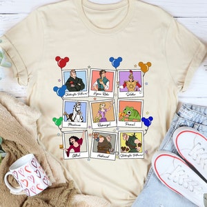 May include: Beige t-shirt featuring a grid of cartoon character portraits in polaroid-style frames. Each frame has a character name below. Colorful Mickey Mouse balloon clusters and stars decorate the design. The shirt is paired with denim shorts and white sneakers.