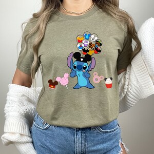 K&ouml;nnte beinhalten: Olivgr&uuml;nes T-Shirt mit einer blauen Zeichentrickfigur mit gro&szlig;en Ohren und einem schwarzen Hut, die Ballons mit Gesichtern von Charakteren h&auml;lt. Das Shirt hat auch Illustrationen eines kandierten Apfels, Zuckerwatte, Donuts und eines Cupcakes.
