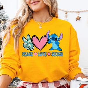 A woman wearing a yellow sweatshirt with a graphic design featuring a blue cartoon character and the text "PEACE LOVE STITCH".