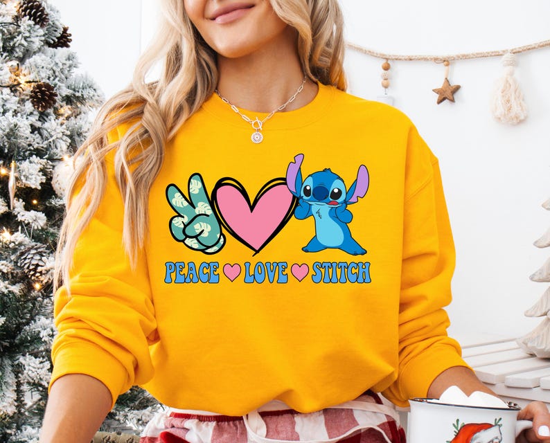 A woman wearing a yellow sweatshirt with a graphic design featuring a blue cartoon character and the text "PEACE LOVE STITCH".