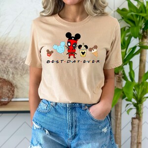 May include: A tan t-shirt with a graphic of a cartoon Deadpool character wearing a Mickey Mouse hat and holding a lightsaber. The graphic also includes Mickey Mouse ears, cotton candy, a Mickey Mouse ice cream cone, and a Mickey Mouse ice cream cup. The text "BEST DAY EVER" is printed below the graphic.