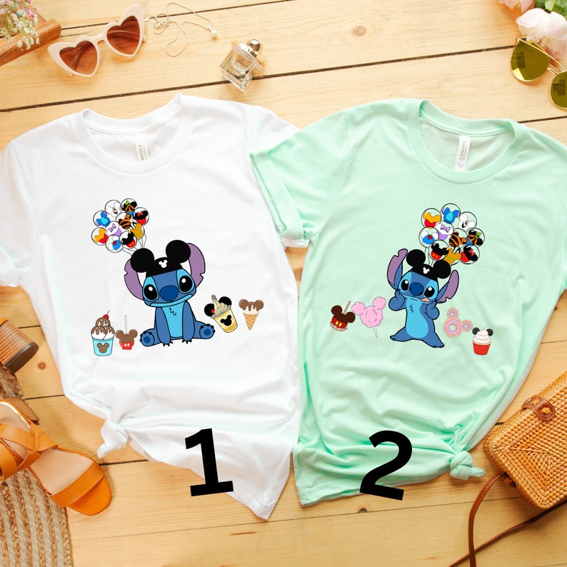 K&ouml;nnte beinhalten: Zwei kurz&auml;rmelige T-Shirts mit Cartoon-Charakter-Designs. Eines ist wei&szlig;, das andere mintgr&uuml;n. Jedes Shirt hat eine Grafik eines blauen Alien-Charakters, der Luftballons h&auml;lt und von Lebensmittelillustrationen umgeben ist. Die Zahlen "1" und "2" sind auf die Shirts gedruckt.