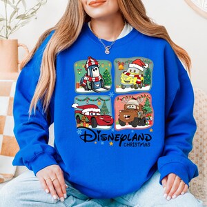 A woman wearing a blue sweatshirt with a collage of four Disney-themed images, including characters from the Cars movie franchise, is sitting on a couch.