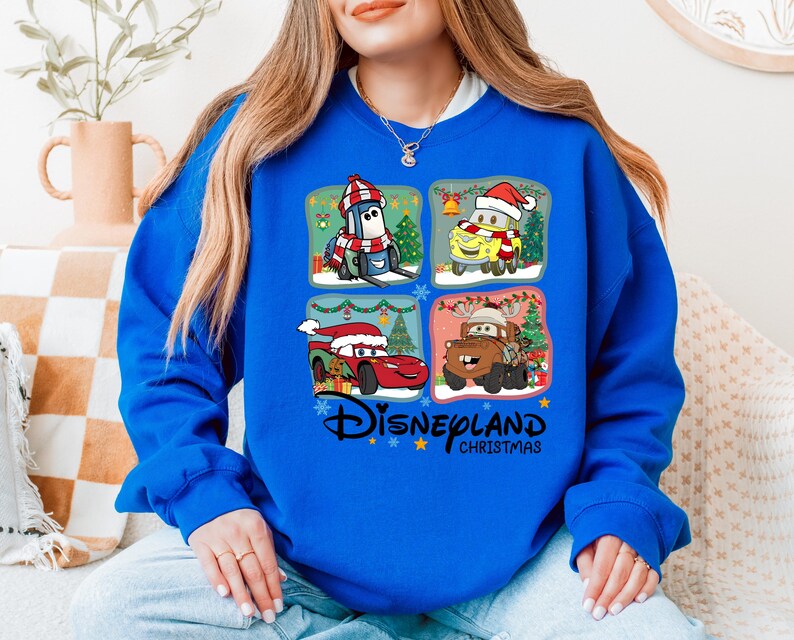 A woman wearing a blue sweatshirt with a collage of four Disney-themed images, including characters from the Cars movie franchise, is sitting on a couch.