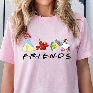 K&ouml;nnte beinhalten: Pinkes T-Shirt mit einem Aufdruck von Arielle, Sebastian, Flounder und Scuttle aus Arielle, die Meerjungfrau. Der Aufdruck ist im Stil des Friends-Logos, mit dem Wort "FRIENDS" unter den Figuren.