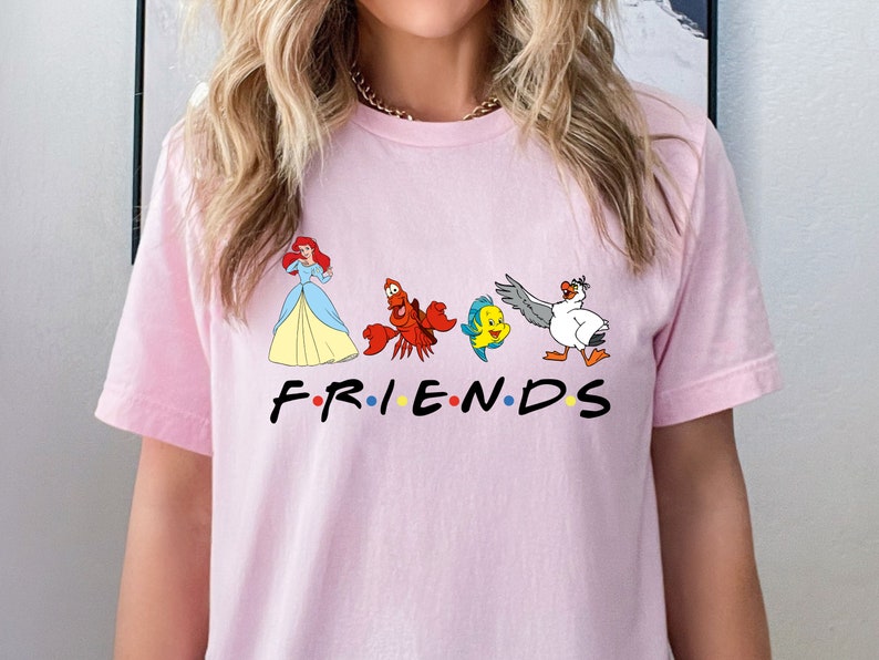 K&ouml;nnte beinhalten: Pinkes T-Shirt mit einem Aufdruck von Arielle, Sebastian, Flounder und Scuttle aus Arielle, die Meerjungfrau. Der Aufdruck ist im Stil des Friends-Logos, mit dem Wort "FRIENDS" unter den Figuren.