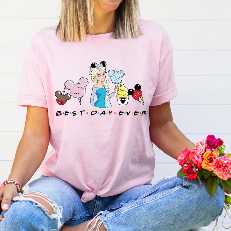 Puede incluir: Camiseta rosa con un gr&aacute;fico de una princesa de Disney sosteniendo orejas de Mickey Mouse, algod&oacute;n de az&uacute;car, un cono de helado de Mickey Mouse y un pretzel de Mickey Mouse. El texto "BEST DAY EVER" est&aacute; impreso debajo del gr&aacute;fico.
