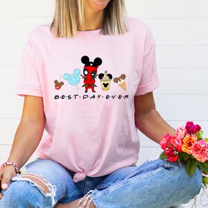 May include: Pink t-shirt with a cartoon image of Deadpool dressed as Mickey Mouse with a cotton candy, Mickey Mouse ice cream cone, and a Mickey Mouse themed drink. The text "BEST DAY EVER" is printed below the image.