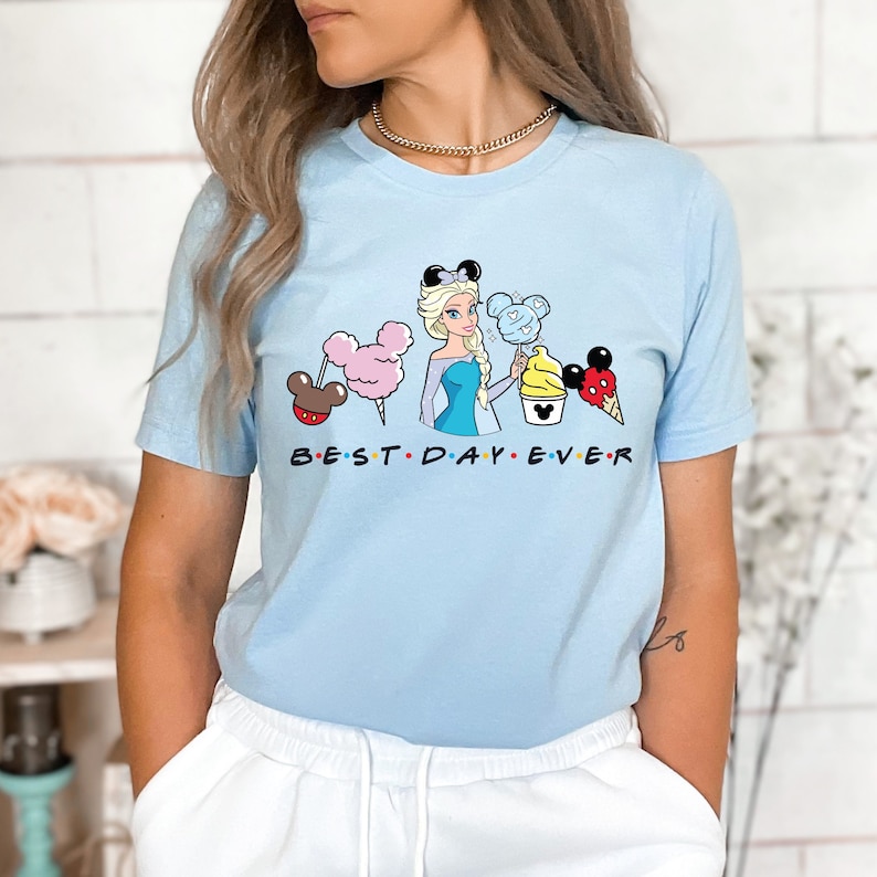 Puede incluir: Camiseta azul claro con un gr&aacute;fico de una princesa Disney de dibujos animados sosteniendo un cono de helado de Mickey Mouse y otras golosinas de Disney. El texto "BEST DAY EVER" est&aacute; impreso debajo del gr&aacute;fico.